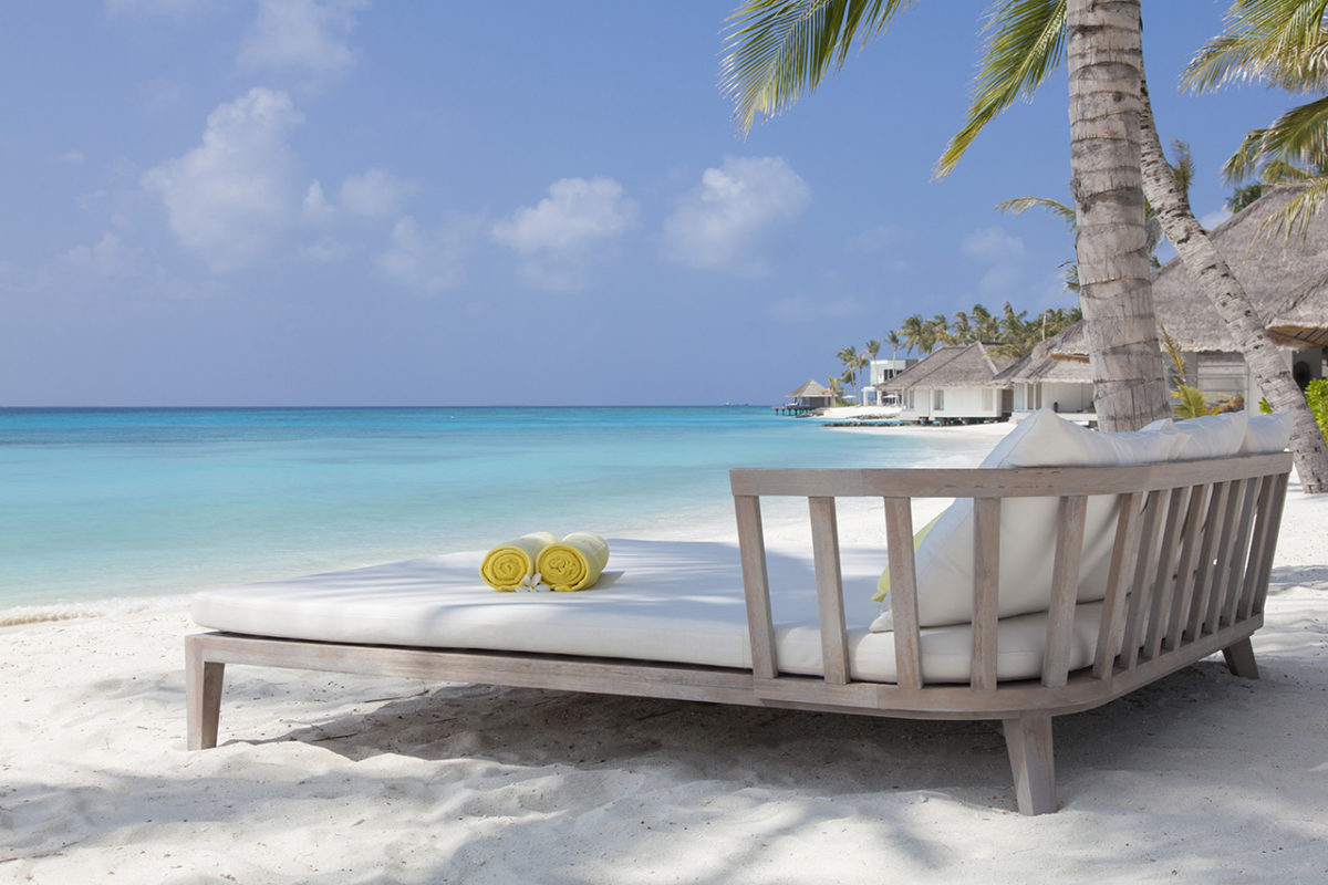 Eine einladenden Liege im schicken Design steht auf dem schneeweißen Sand des Luxusresort Cheval Blanc Randheli auf den Malediven und eröffnet den Blick auf die türkisfarbene Lagune des Luxusresort