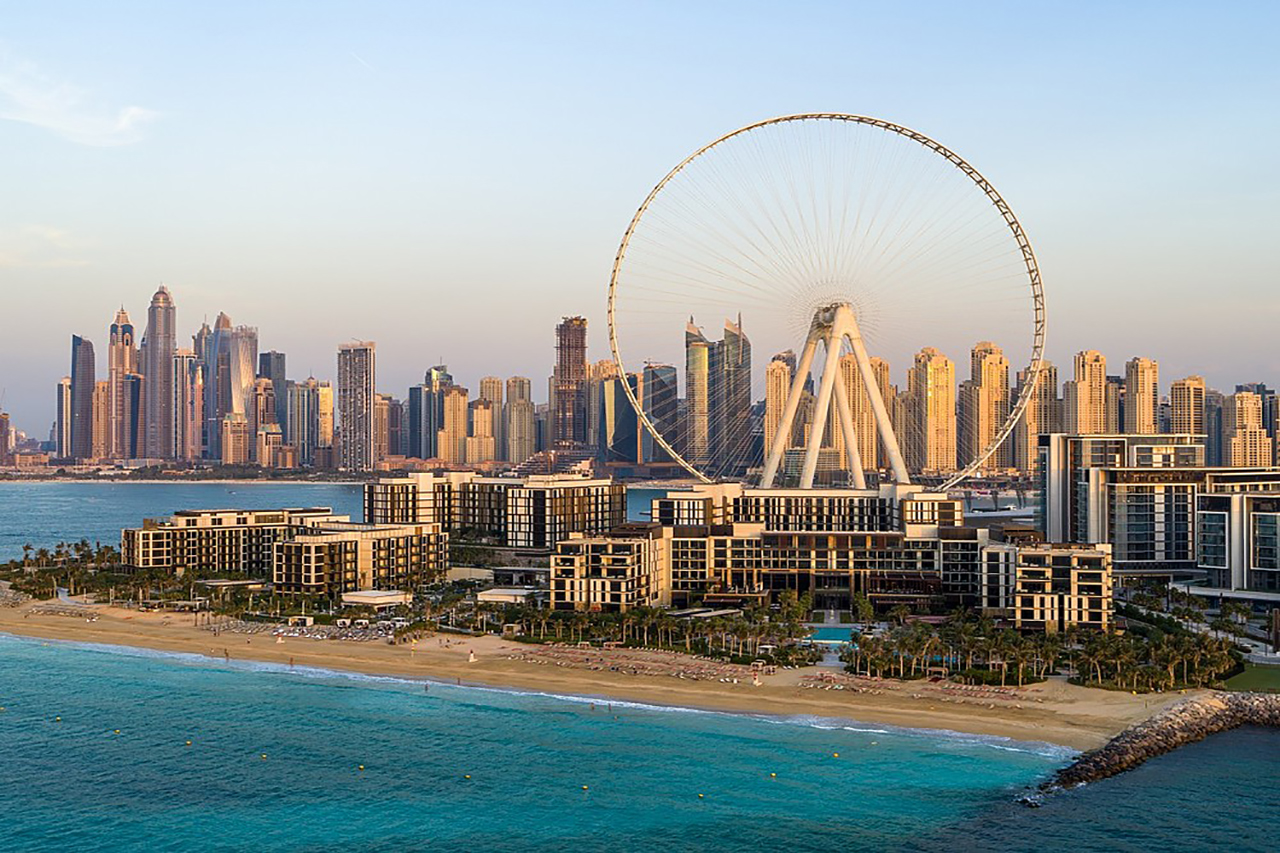 Blick vom Wasser aus auf das Caesars Palace Bluewater Dubai vor dem Jumeirah Beach mit dem Cove Bay Club rechts daneben und dem Riesenrad im Hintergrund bei Sonnenschein in Dubai